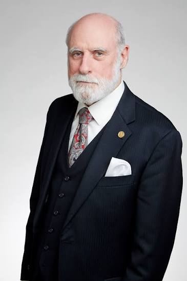 Homem idoso de pele clara, calvo no topo da cabeça, possui cabelos brancos nas laterais e barba grisalha. Ele usa óculos de grau e veste um elegante terno cinza com colete.