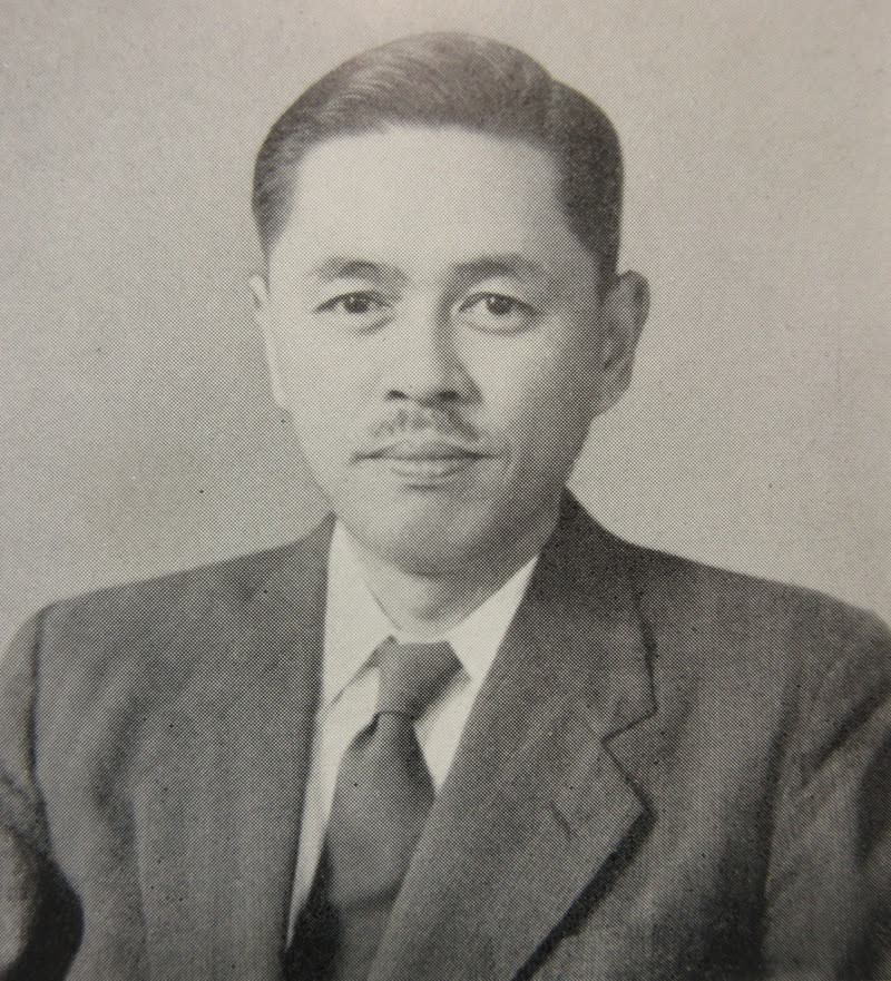 Fotografia em preto e branco de homem japonês de meia-idade, cabelos escuros penteados para trás e bigode fino. Veste terno escuro, camisa clara e gravata.