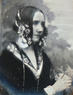 Retrato de época, em tons sépia, de mulher jovem de cabelos escuros repartidos ao meio e presos em cachos laterais adornados com flores. Veste vestido escuro de gala com decote adornado e colar, mãos cruzadas à frente do corpo, sobre um fundo com nuvens.