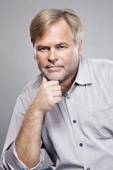 Homem branco de pele clara, com cabelos castanho-claros levemente grisalhos e volumosos, divididos ao meio. Possui olhos claros, formato de rosto arredondado, expressão pensativa com a mão apoiada no queixo, barba curta e camisa cinza-clara.