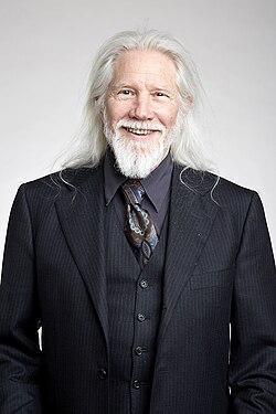Homem branco, com longos cabelos brancos e farta barba grisalha. Olhos claros, expressão serena e amigável, terno elegante e bem alinhado.